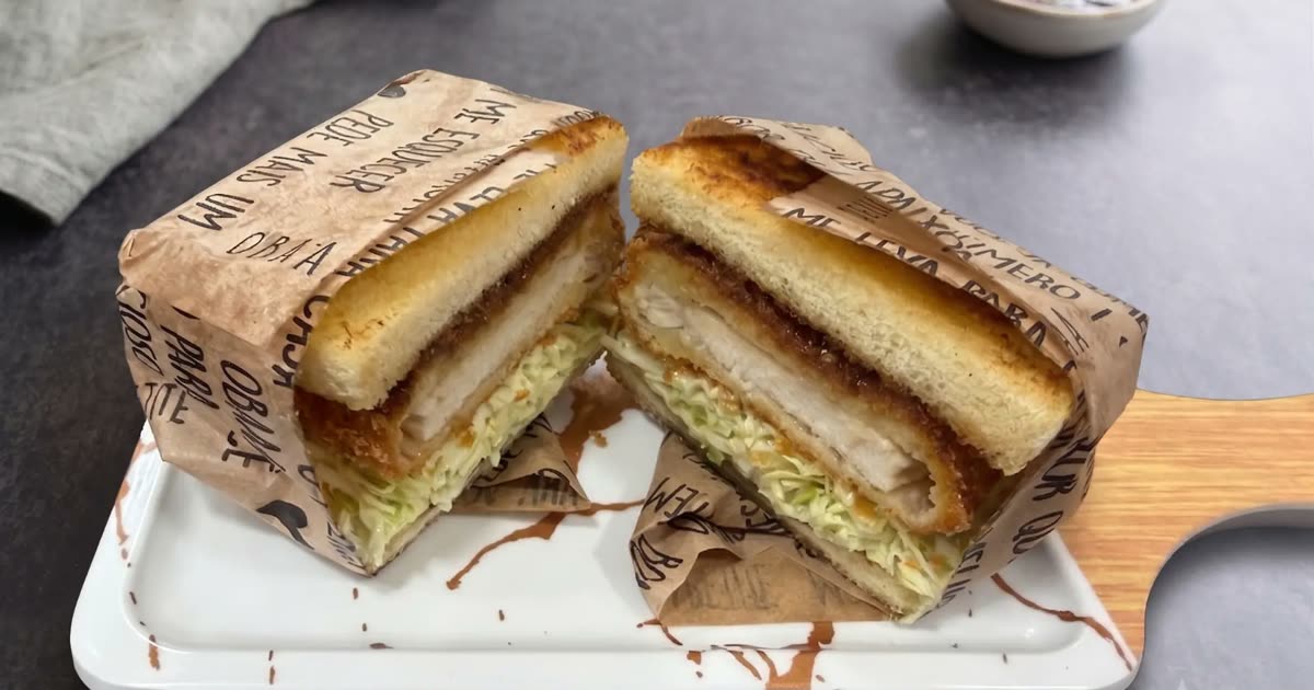 Sando japonês de frango empanado cortado ao meio, embrulhado em papel, sobre tábua de madeira